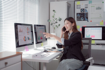 Businesswoman is sitting at her desk, analyzing financial charts on her computer and talking on her mobile phone, discussing business strategy and market trends