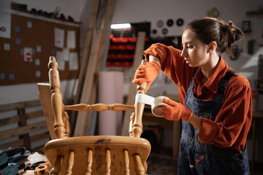 repair and diy concept. Young woman in her dark workshop sticking adhesive tape to old wooden chair