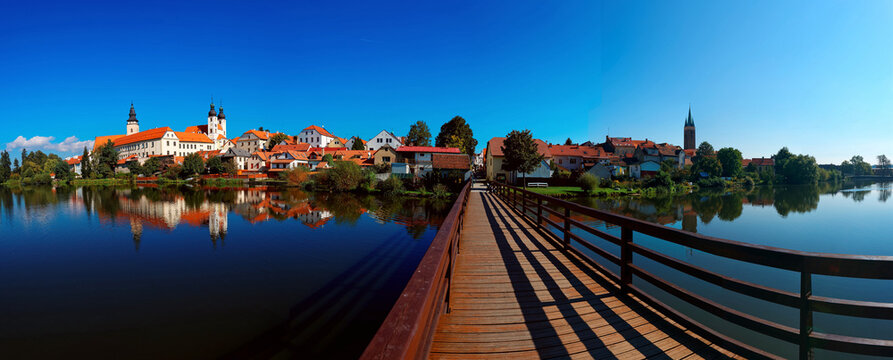 Wide panoramic view of beautiful Telc old town by lakeside, with red-roof houses reflected in the peaceful water & a bridge under blue sunny sky~ A UNESCO world heritage town in Czech Republic, Europe