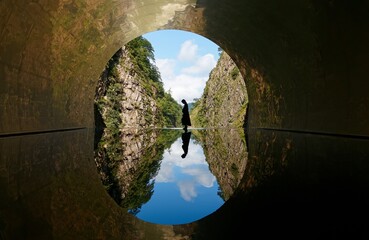 A tourist standing at the end of a tunnel with the inverted landscape of Kiyotsukyo Gorge reflected...