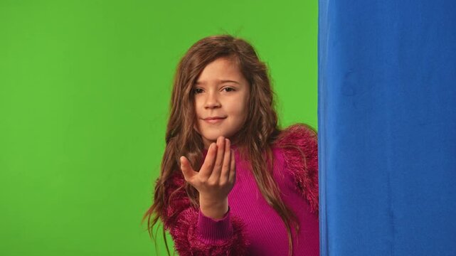 Little girl child peeks out waves hand and shows come here gesture, positive expression, isolated on chroma key green screen studio background mockup.