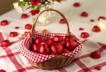 An overhead photo of a picnic setup featuring a red and white checkered blanket, with heart-shaped candies scattered around a basket filled with treats. The scene includes a bouquet of fresh flowers a