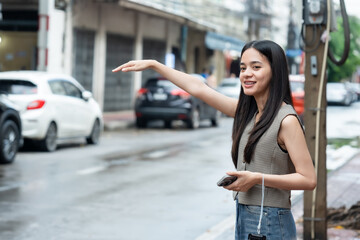 Fototapeta premium Beautiful Asian tourist walking in old city of Bangkok. Smiling young woman with long black hair waving hand to hail taxi. Holding smartphone and wearing stylish vest with casual jeans.
