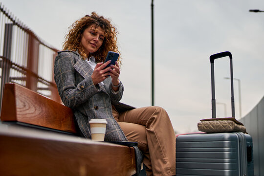 Sitting on a bench, a Caucasian woman uses her phone, holding a takeaway coffee with a suitcase beside her, patiently waiting for the bus.