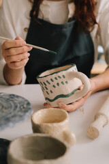 Experienced artist painting designs on ceramic cup in pottery workshop. Potter draws an abstract design into earthenware cup.