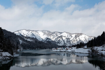 Beautiful scenery of snowy mountains reflected in the peaceful water of River Tadami Gawa and a village perched on the riverside slope on a sunny, cloudy winter day in Oku-Aizu, Fukushima, Japan