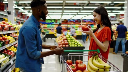 Multiethnic couple selecting fresh produce, walking through well stocked supermarket aisle, sharing moments while choosing fruits, pushing shopping cart in smooth slow motion sequence