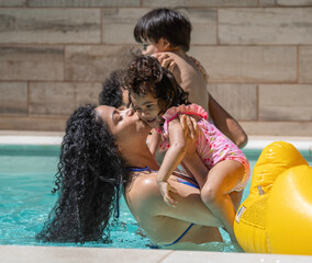 Happy latin family enjoying summer vacations in a swimming pool and mother is kissing her daughter