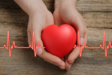 Cardiology. Woman holding red decorative heart on wooden table, closeup. Red heartbeat line