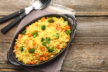Tasty rice with vegetables in baking dish and cutlery on wooden table, top view. Space for text