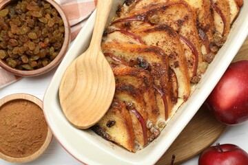 Delicious bread pudding in baking dish, fresh apples, cinnamon and raisins on white table, flat lay