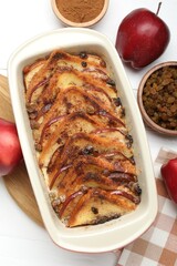 Delicious bread pudding in baking dish, fresh apples, cinnamon and raisins on white table, flat lay