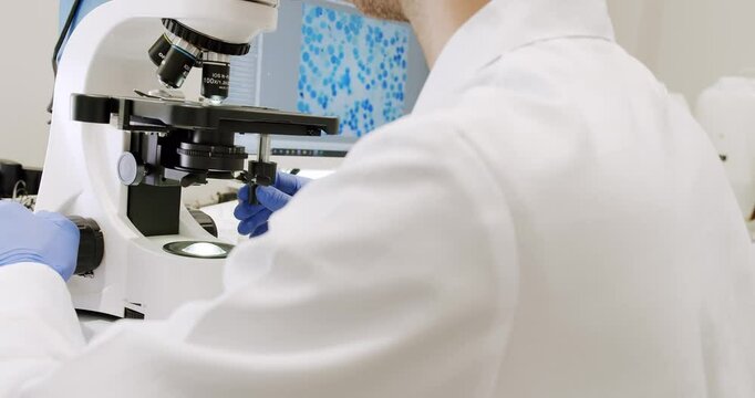 Man in lab coat and latex gloves using microscope to examine sample during work in clinic in daytime. Using a microscope to examine the patients blood health condition.Microscope for blood examination