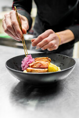 A chef delicately garnishes a black bowl with sliced chicken, pickled onions, and lemon, showcasing culinary skill and attention to detail in a modern kitchen setting.