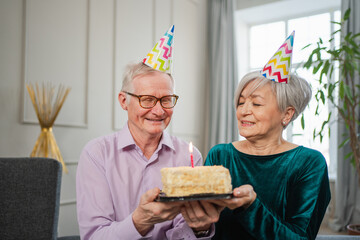 Make a wish. Family senior couple in party hat cap celebrating birthday anniversary together at home. Old man blowing out burning candles on birthday cake. Old woman wishes husband happy birthday
