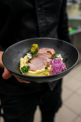 A chef presents a gourmet dish featuring tender slices of meat atop creamy mashed potatoes, garnished with pickled vegetables and fresh herbs in a stylish black bowl.