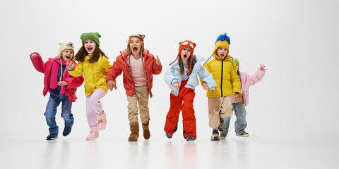 Group of children, dressed colorful winter jackets and hats, running forward with excited expressions, showing joy and energy against white studio background. Concept of childhood, winter vacation