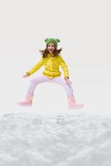 Little girl wearing red jacket, beige pants, winter boots jumping high, arms outstretched, smiling joyfully, celebrating winter fun against white background. Concept of fashion, winter vacation.