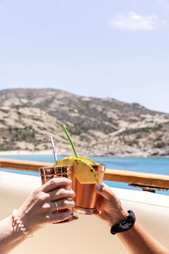 Two female hands holding cocktails in copper cups on a yacht in Greece toasting on their sailing holiday in the summer. 