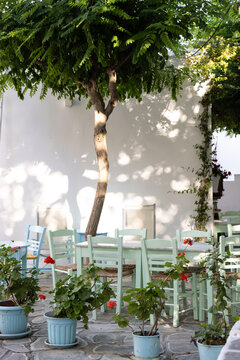 Quiet scene in a small Greek village with leafy trees, colorful tables and chairs, and dappled sunlight. 