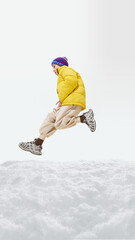 Boy in yellow puffer jacket, blue hat leaping with one leg forward, arms tight, winter adventure in...