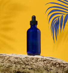 Blue glass bottle with black pipette and palm branch on yellow background, oil and cosmetics