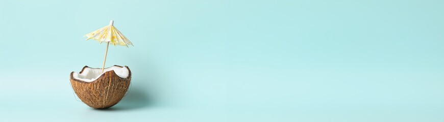 Half coconut shell with a yellow cocktail umbrella on a pastel turquoise background. Minimalist studio food photography. Summer vacation and tropical drinks concept.