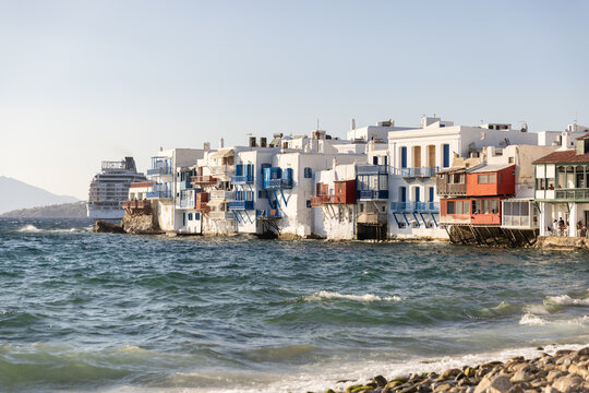 Horizontal images of red, white, and blue buildings in Mykonos, Greece in late afternoon with waves crashing on a pebbled shore. 