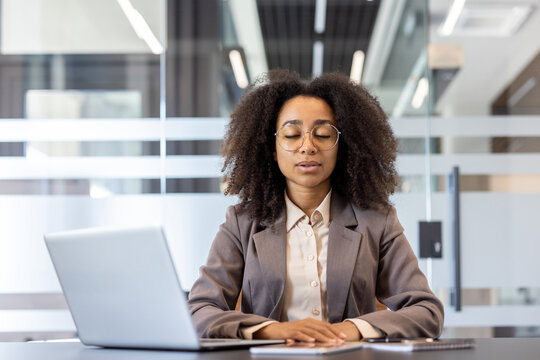 Young african american business woman in suit sitting at desk in office, resting eyes closed and breathing deeply