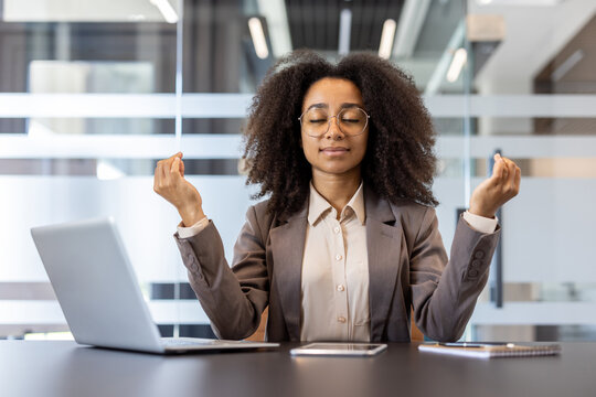 Relaxed young African American woman in suit sitting in office at desk in lotus position with closed eyes, meditating and relaxing