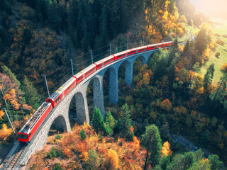 Aerial view of modern red train on Landwasser viaduct in alpine mountains, orange forest at sunset...