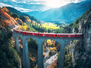 Aerial view of modern red train on Landwasser viaduct in alpine mountains, orange forest at sunset...