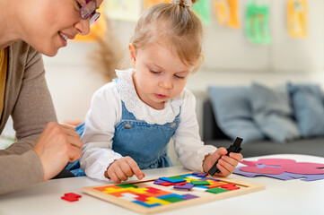 A caring teacher and a toddler girl engage in a hands-on learning activity with colorful wooden...