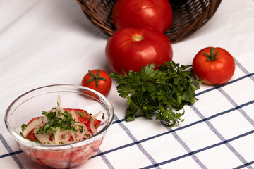 ensalada chilena, un bol de vidrio con tomate, cebolla y perejil picado, al fondo un canasto con tomates maduros y ramas de perejil