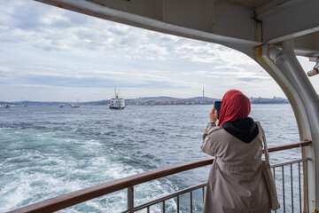 Muslim woman capturing scenic view of Istanbul's Bosphorus from ferry ride with city skyline in background. Enjoying the ferry ride across Istanbul's Bosphorus Strait. Headscarf woman taking pictures