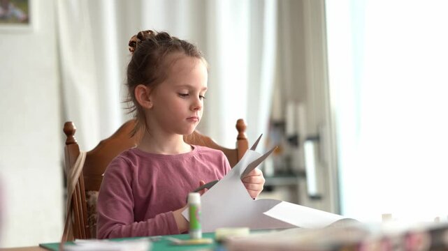 Cute little girl with her hair up cuts paper with scissors while sitting at the table. Children's creativity and education concept
