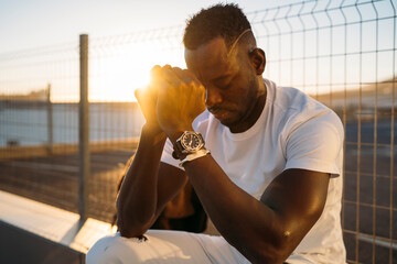Young african american man praying with hands together at golden hour near fence
