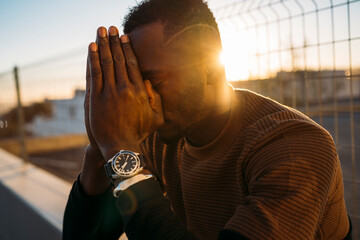 Young african american man praying with hands on face at sunset