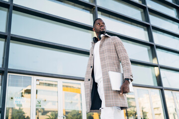 Stylish entrepreneur walking with laptop in hand near modern office building