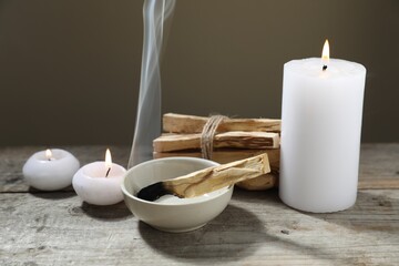 Smoldering palo santo stick and burning candles on wooden table against grey background, closeup