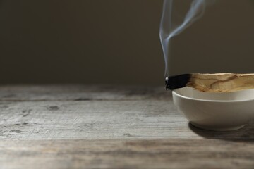 Smoldering palo santo stick and bowl on wooden table against grey background, closeup. Space for text