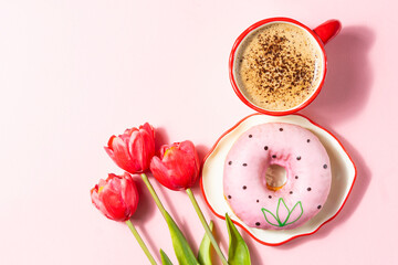 A cup of coffee with a pink donut and red tulips on a pastel pink background. A delicate and festive composition for Women's Day or a spring celebration.
