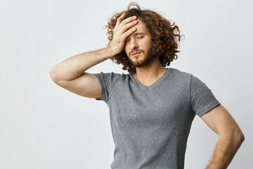 A frustrated man with curly hair in a gray t shirt, holding his forehead, conveying stress against...