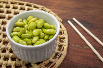 close-up, of a small, white bowl filled with edamame green soy beans, bowl lies on a straw mat on a wooden surface.