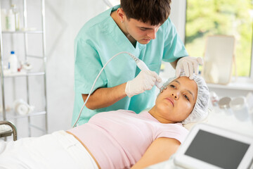 Fototapeta premium Young male cosmetologist performs hardware facial microcurrent procedure to young female patient