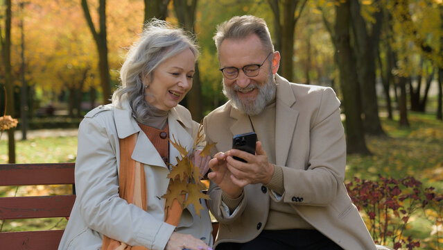 Caucasian retired couple family in love mature woman man sit on bench autumn park using mobile phone gadget together smiling wife husband spouses reading news old age and smartphone in city outdoors