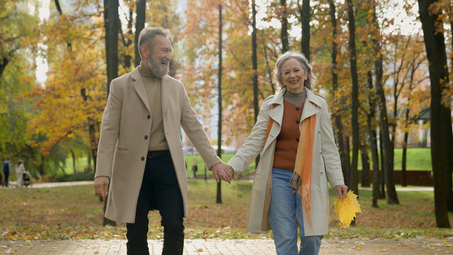 Middle aged 60s couple gray-haired wife and husband in elegant outerwear walking romantic date in autumn city park Caucasian happy family woman man grandparents holding hands together walk outdoors