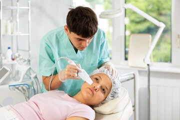 Young male cosmetologist performs hardware facial exfoliating procedure to young female patient
