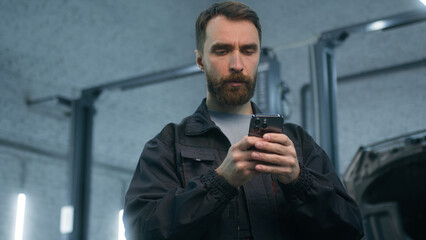 Mechanic man auto mechanical worker using mobile phone gadget in repair car service garage automobile factory pensive thoughtful male guy repairman look at smartphone screen think typing on cellphone