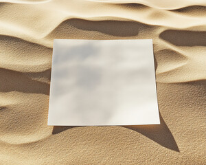Fototapeta premium A blank white sheet of paper placed on a sandy surface with soft shadows, symbolizing space for creativity, design, and text, ideal for mockups and advertising.